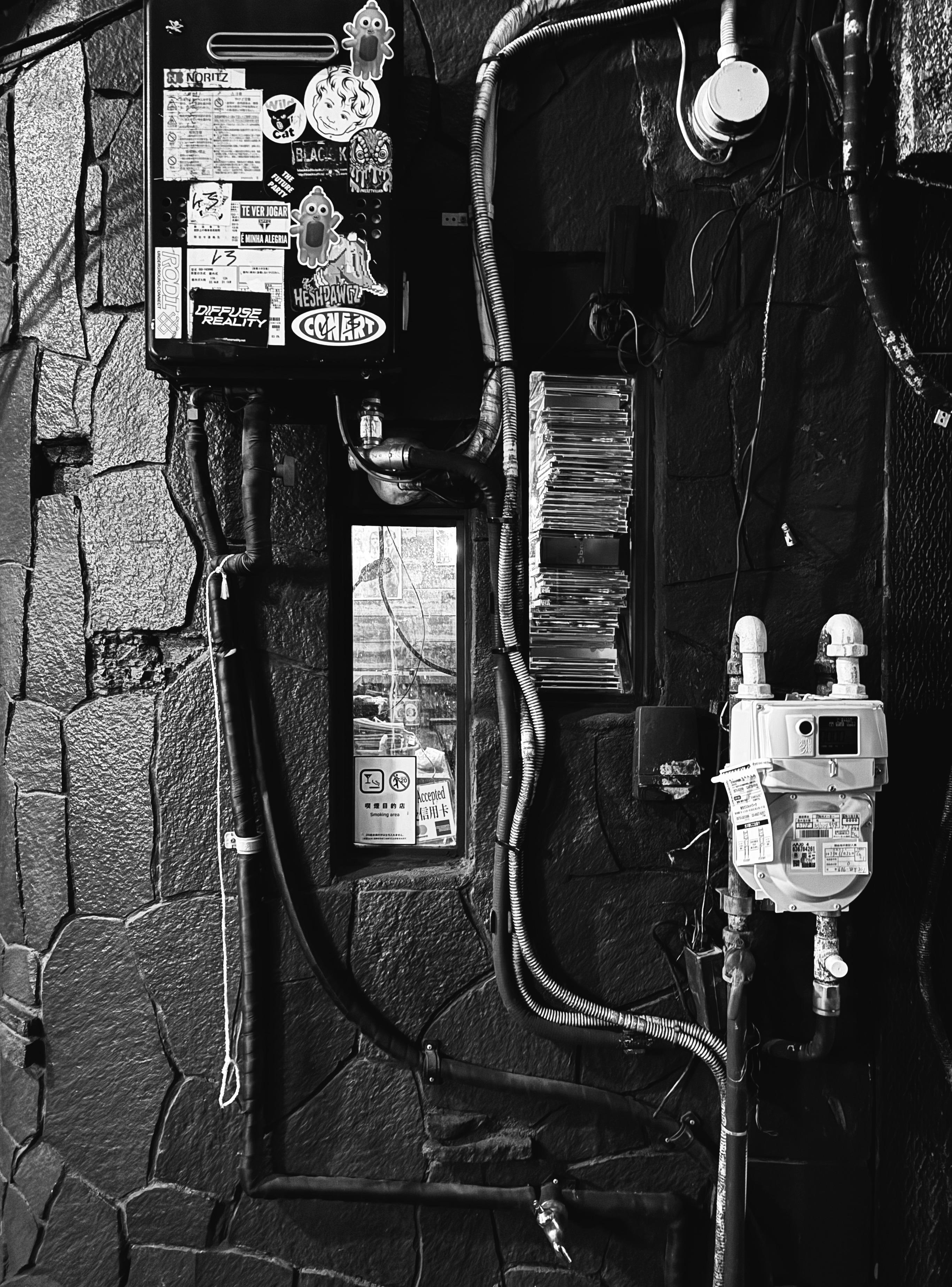 Black and white photo of a brick wall and a tiny window, wires running to a meter, stickers all over a meter