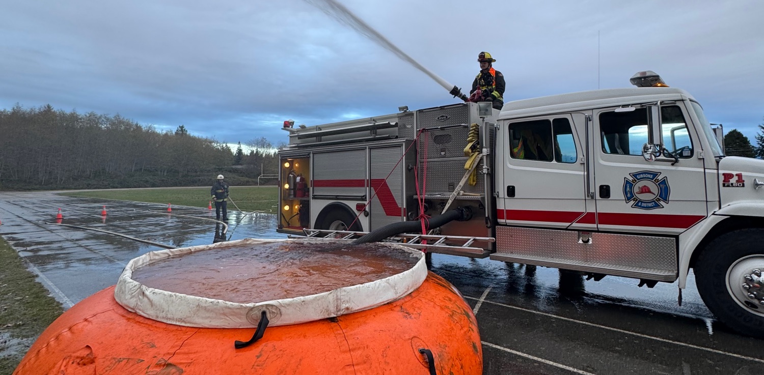 A firefighter sits atop a fire engine spraying water from a monitor, the truck is being supplied via drafting from an orange pumpkin tank