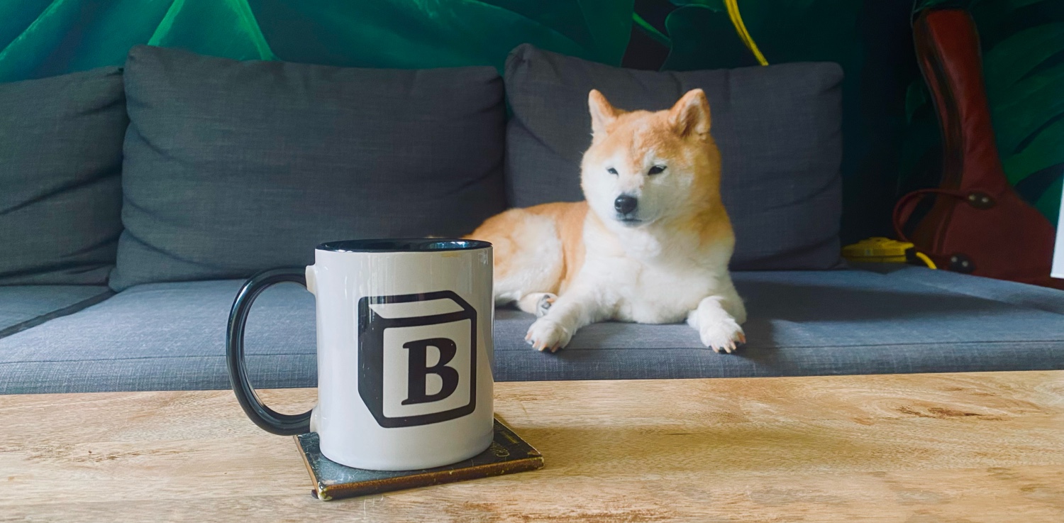 Photo of a coffee mug with a 'B' on it sitting on a table in front of a Shiba Inu (dog)